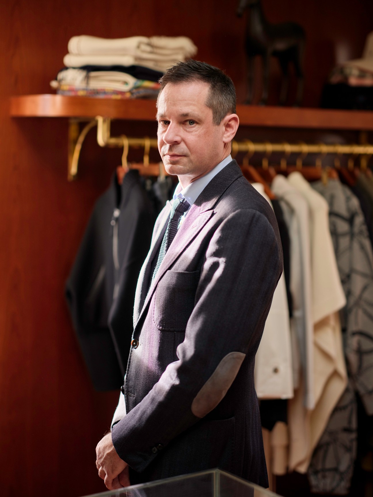 Man in dark blazer and pink shirt standing in Cuffs boutique with clothing displayed on wall behind