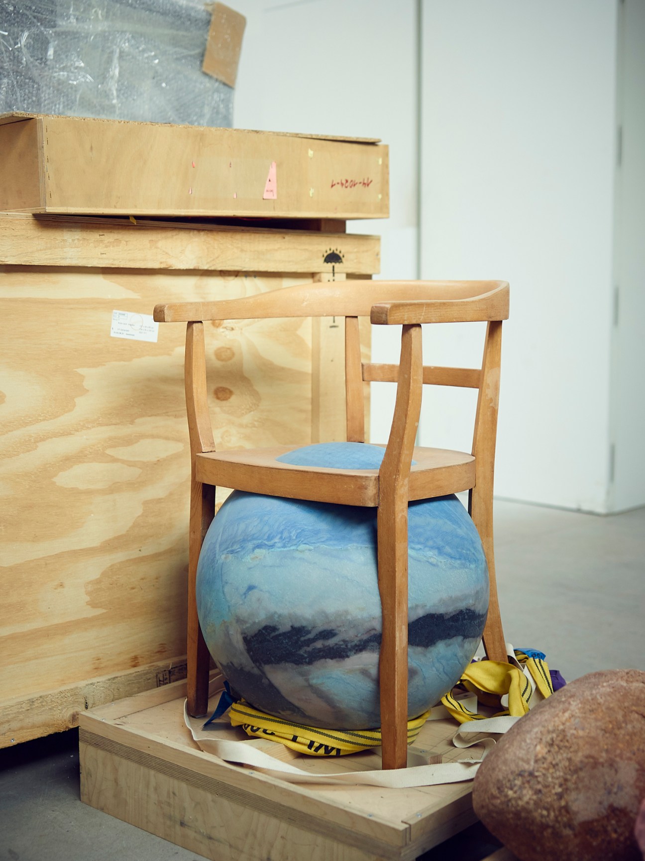 Wooden chair with large blue spherical sculpture nestled in seat among wooden crates and materials