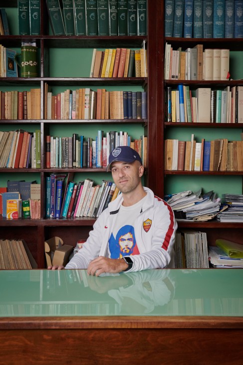 Vincenzo Belpiede sits at a desk in front of a wooden bookshelf filled with books, wearing a navy cap and light-colored jacket.