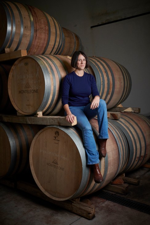 Giulia Monteleone sits on wine barrels marked with her vineyard name in a cellar.