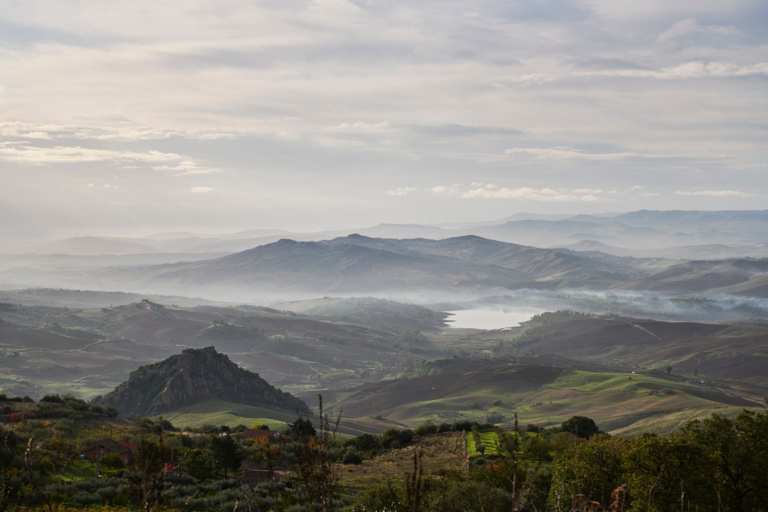 Rolling hills and valleys of Sicily's remote interior with misty mountains in the distance and a lake nestled between green and brown farmland.