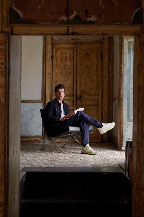 Dario Nepoti sits reading in a worn interior space with aged wooden doors and weathered walls.