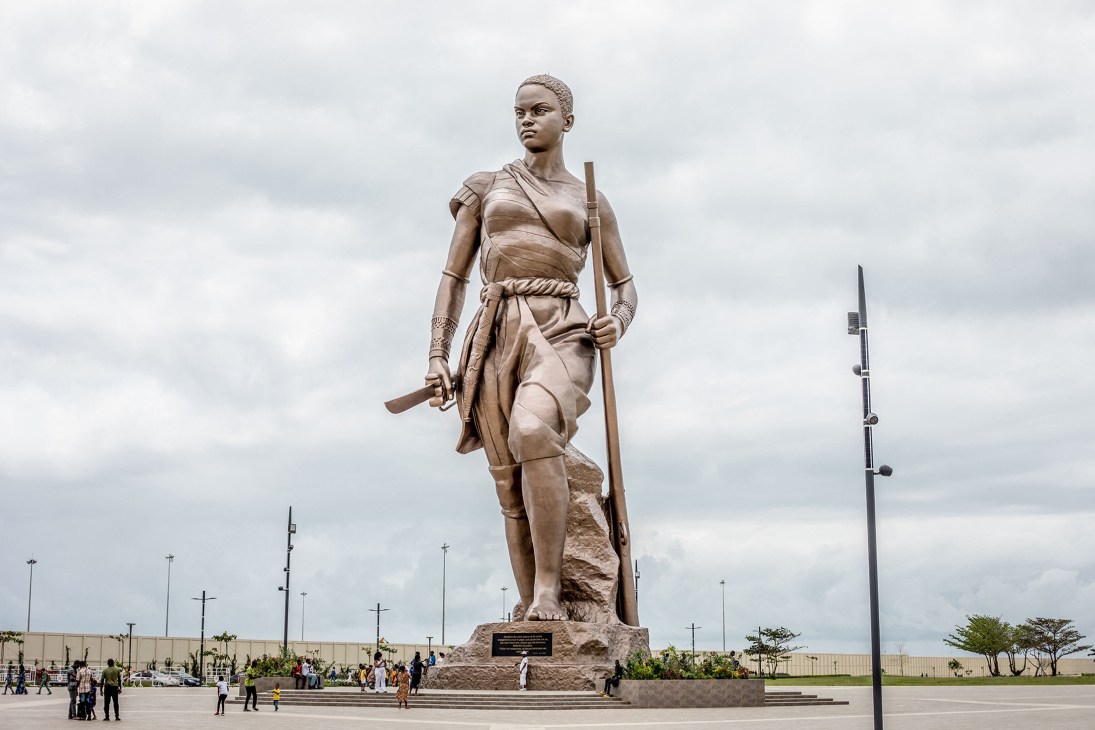 Large bronze statue of a female warrior figure holding a staff against cloudy sky