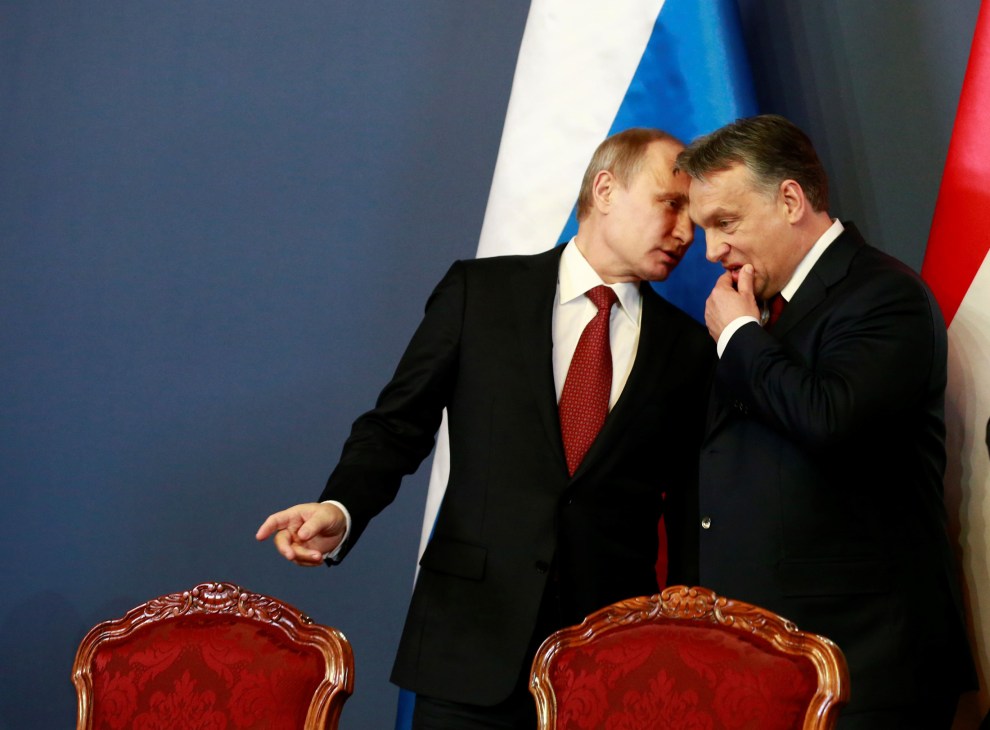 Vladimir Putin and Viktor Orbán leaning close together in conversation, with ornate red chairs and flags in background