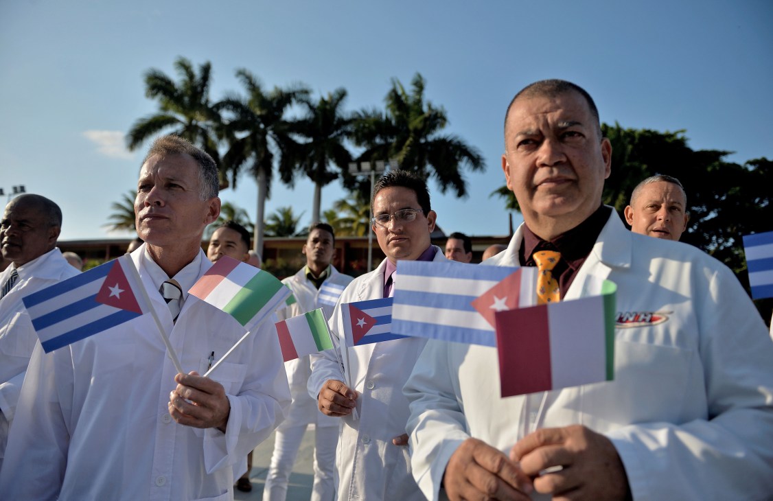Cuban medical professionals in white coats holding small Cuban flags at an outdoor ceremony