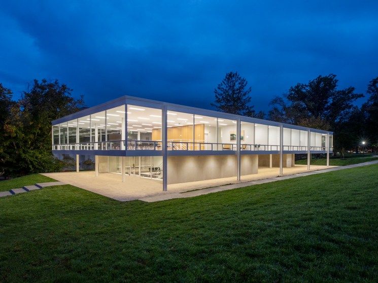 Modern glass and steel building with floor-to-ceiling windows illuminated at dusk surrounded by green lawn