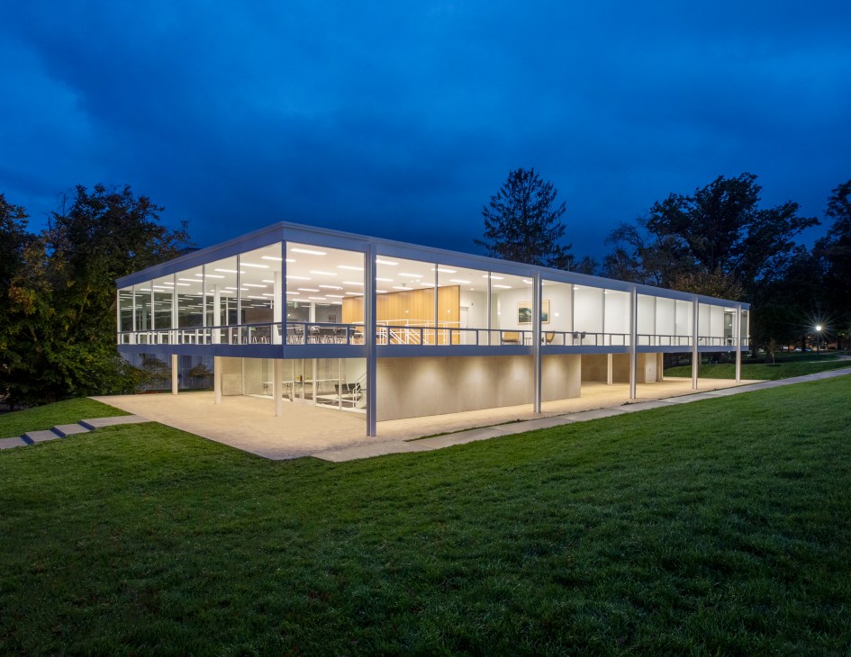 Modern glass and steel building with floor-to-ceiling windows illuminated at dusk surrounded by green lawn