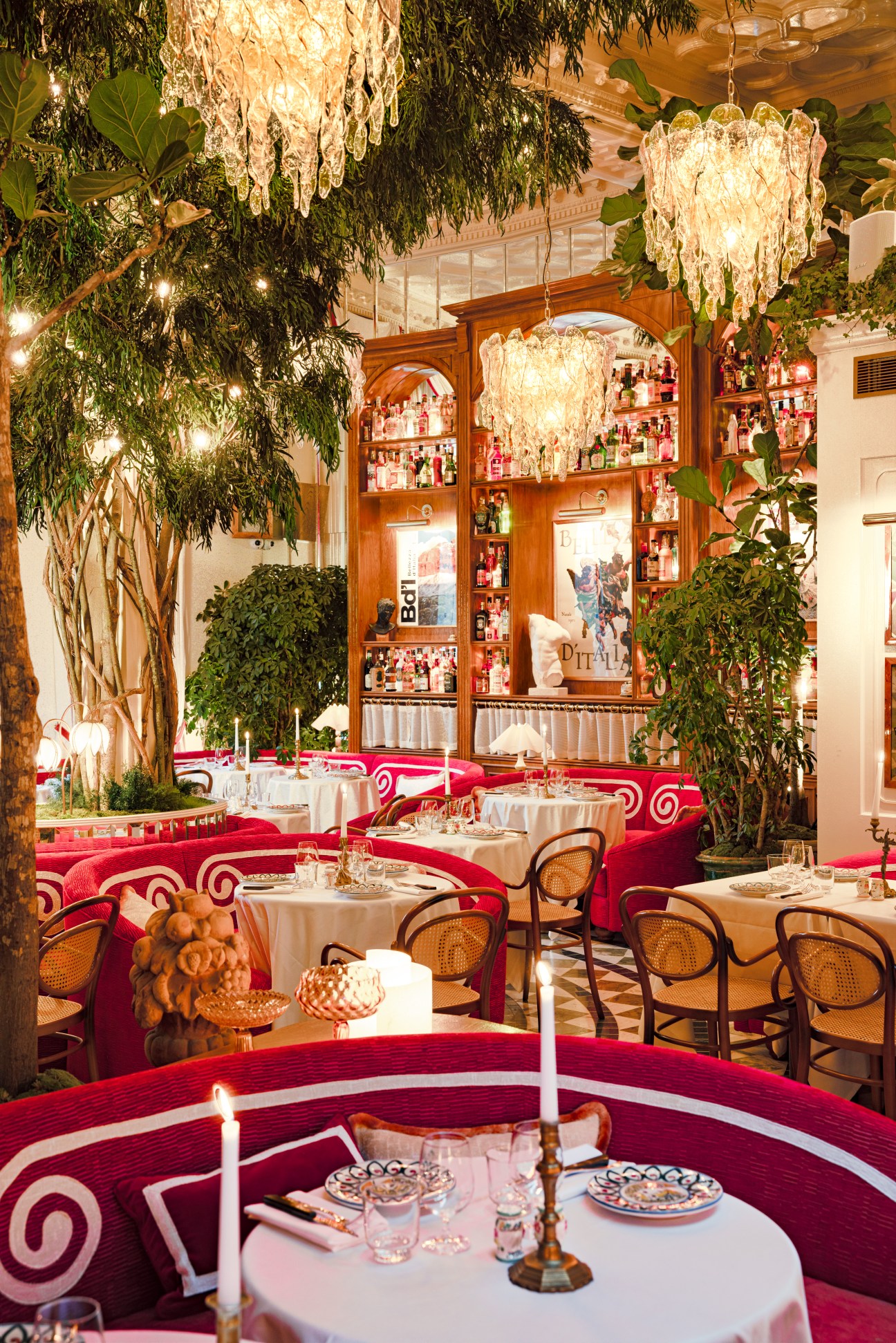 Restaurant dining room with crystal chandeliers, red velvet banquettes, potted plants and formal table settings