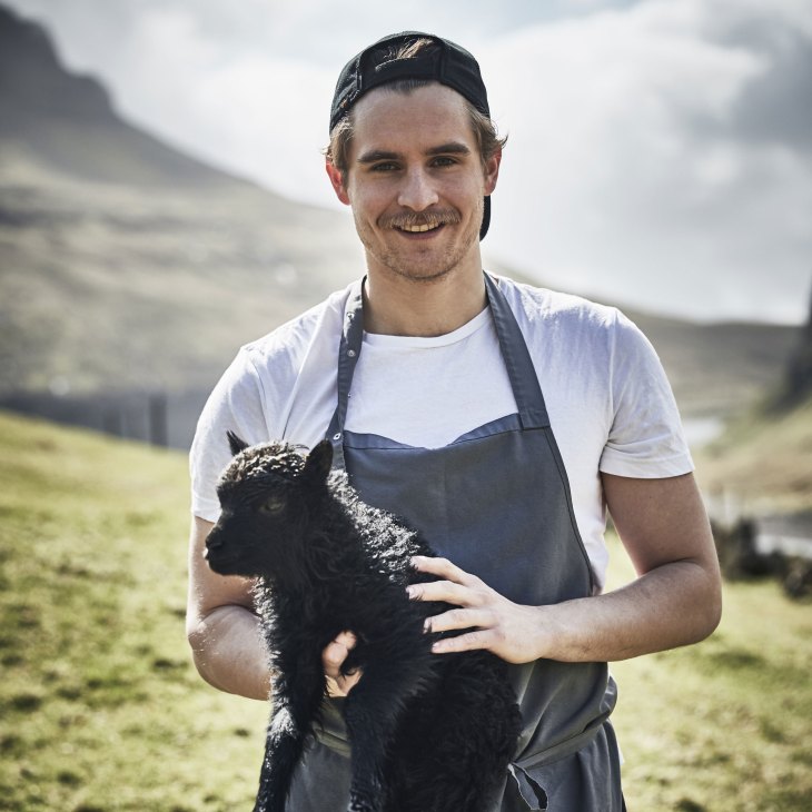 Chef working with black lamb in outdoor mountain setting wearing apron and cap