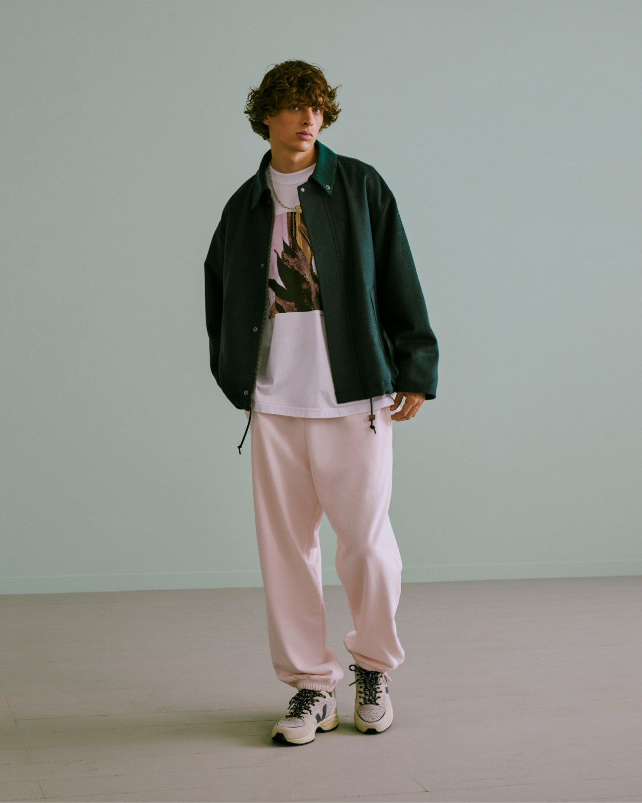 Young male model in curly hair wearing dark green jacket over light pink shirt and pink sweatpants from Citen collection