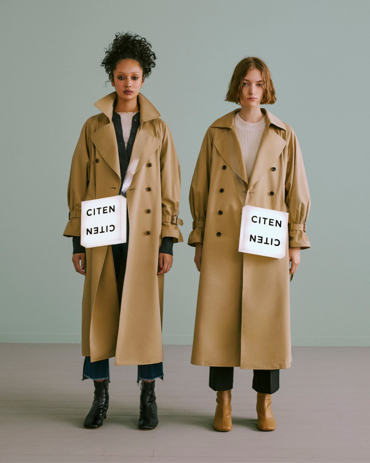 Two women wearing beige trench coats holding white bags with 'CITEN' branding in a studio setting