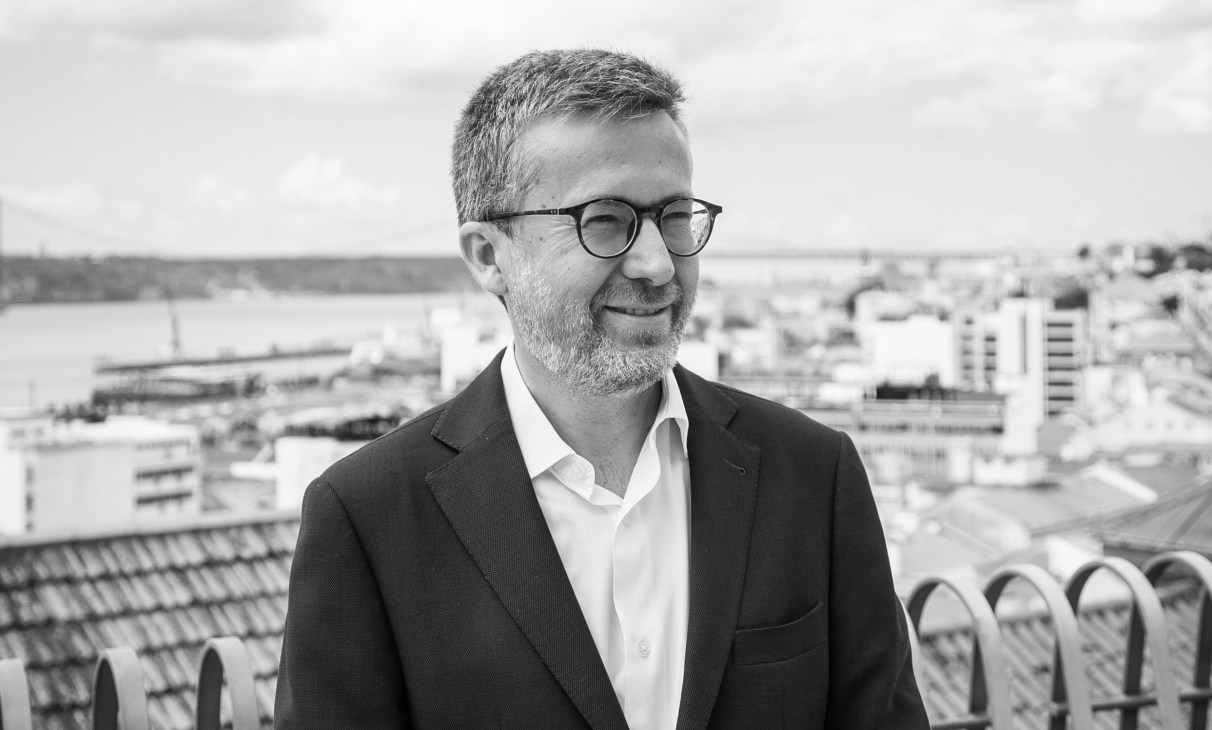 Man wearing glasses and dark blazer stands on rooftop overlooking Lisbon cityscape with river and buildings in background