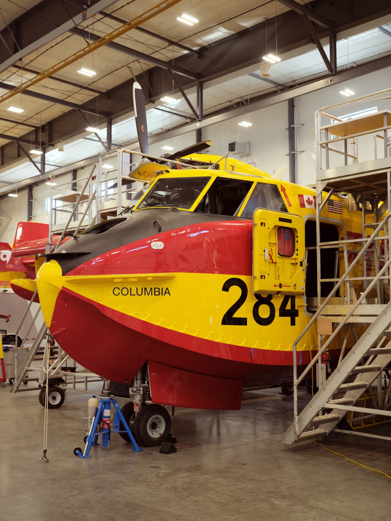 Yellow and red firefighting aircraft with number 284 and 'COLUMBIA' text in maintenance hangar with access stairs