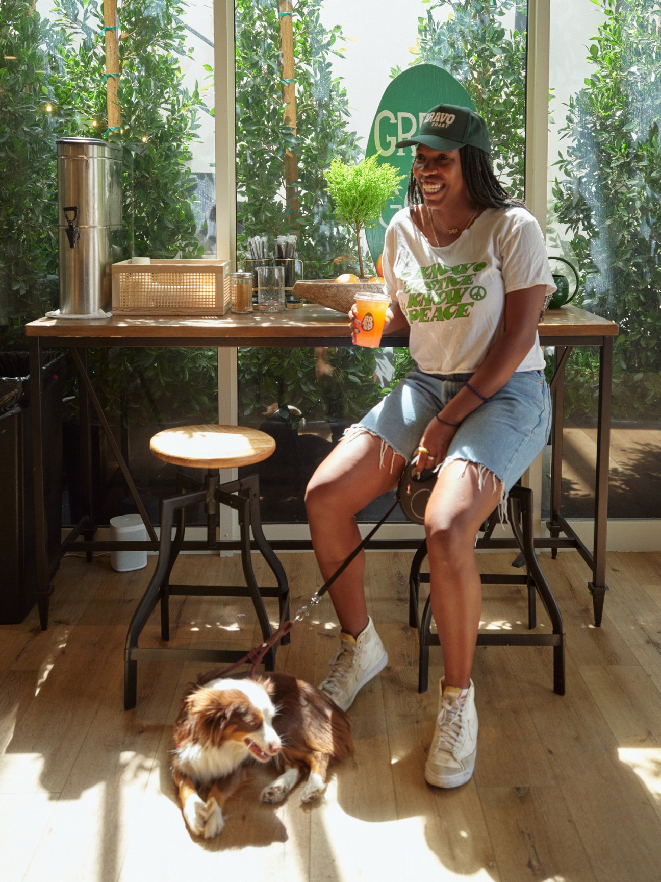 Person in green cap and white t-shirt sitting at counter with drink, small dog on leash below in bright plant-filled cafe