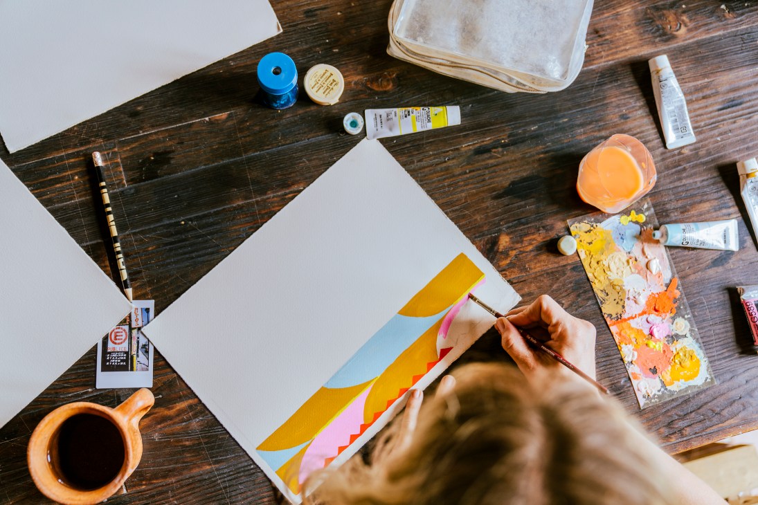 Colorful art materials and painting supplies scattered on wooden table with hands holding brush