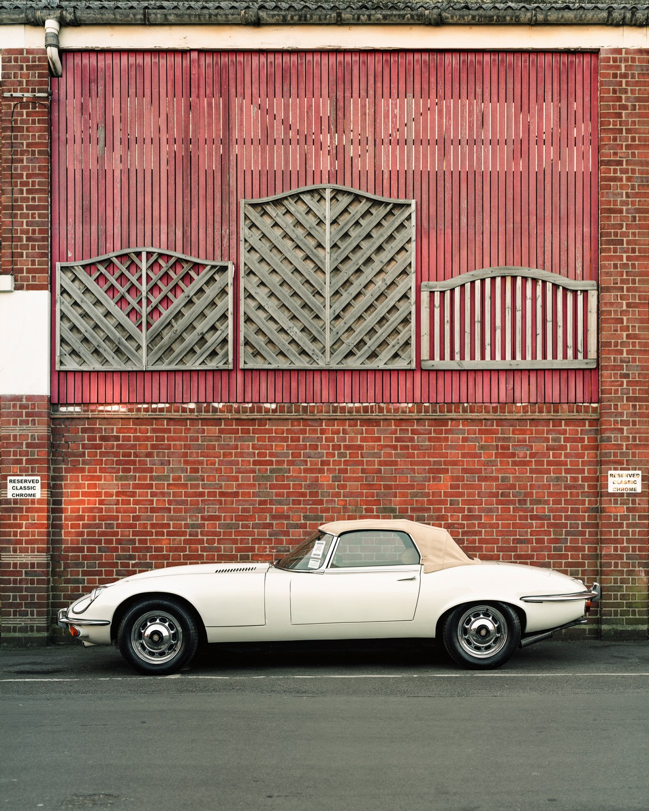 White Jaguar E-Type Series 3 convertible with soft top parked against brick wall with decorative fence panels