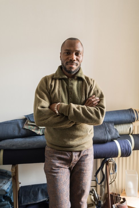 Portrait of Marly Manku, French tailor, standing with arms crossed in front of rolled fabric bolts at La Clinique du Jean.