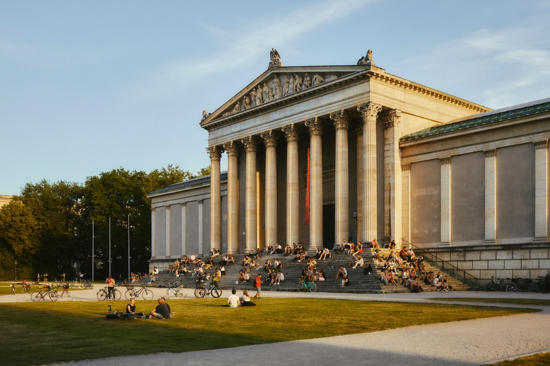 Classical building with columns and people sitting on steps in Munich