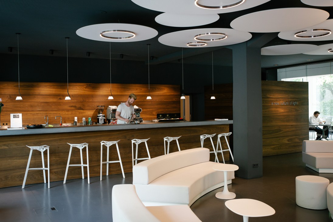 Modern office interior with coffee bar, white bar stools, circular ceiling lights, and curved white seating area