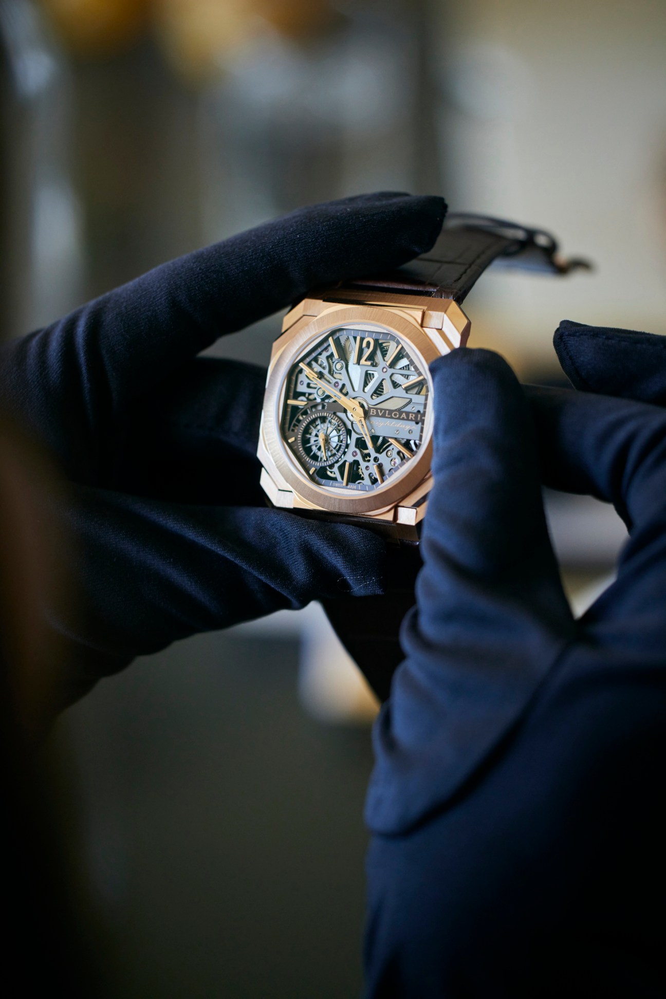 Hands wearing dark gloves carefully holding a luxury Bulgari watch with skeletal dial and rose gold case