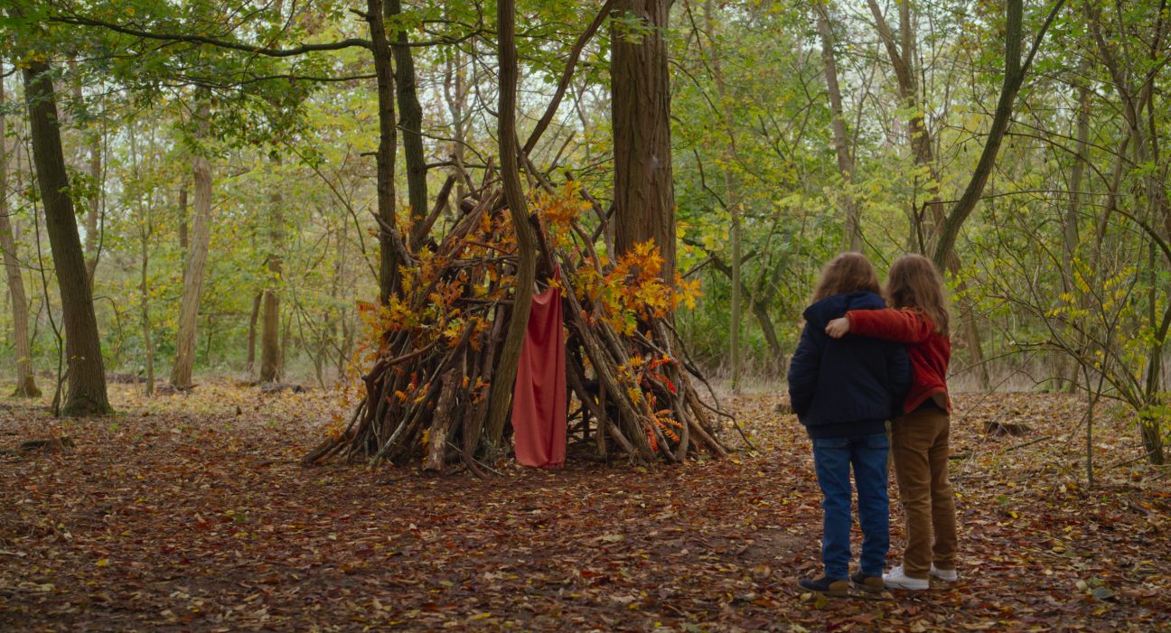 Two children embracing while looking at a makeshift fort built from branches and decorated with autumn foliage