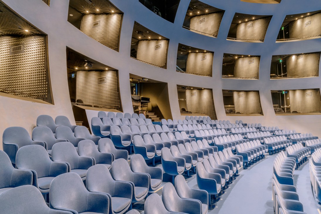 Modern auditorium with rows of blue-grey seats and geometric golden acoustic panels in ceiling alcoves