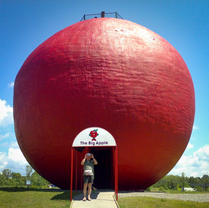 A visitor stands at the entrance of The Big Apple, a giant red apple sculpture with cartoon eyes and a smile in Colborne, Ontario.