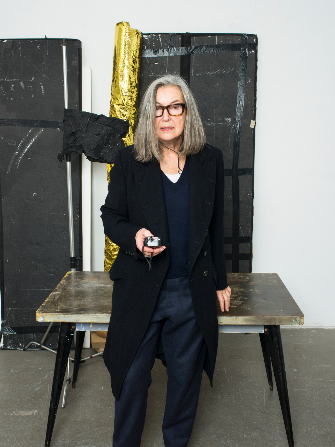 Photographer Elfie Semotan in her Vienna studio, holding a vintage camera, standing in front of black and gold abstract artwork.