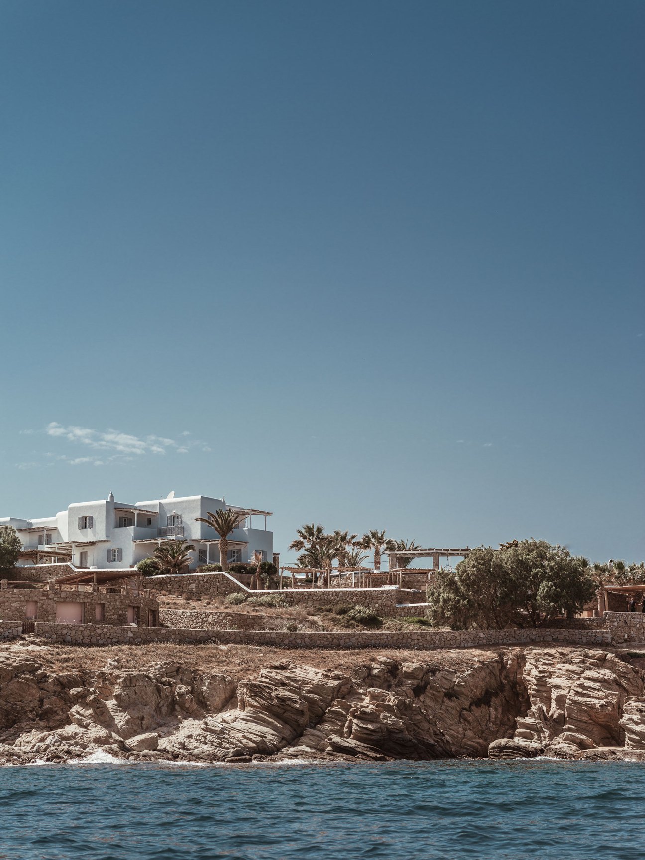 White Cycladic-style building with palm trees and wooden pergolas overlooking rocky coastline and blue sea at Soho Roc House, Mykonos.