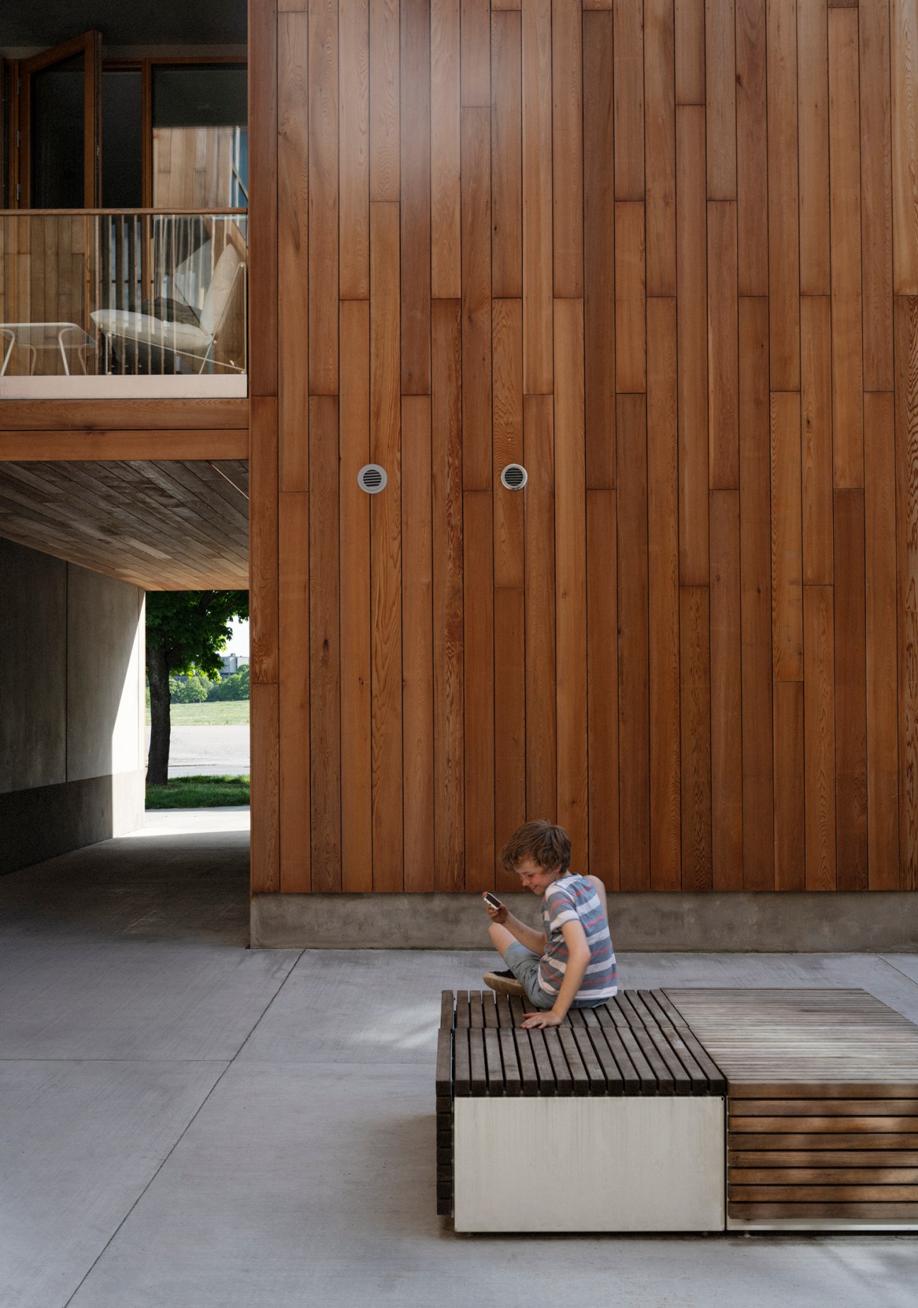 Child sitting on a wooden bench in a modern courtyard surrounded by cedar-wrapped wooden architecture.