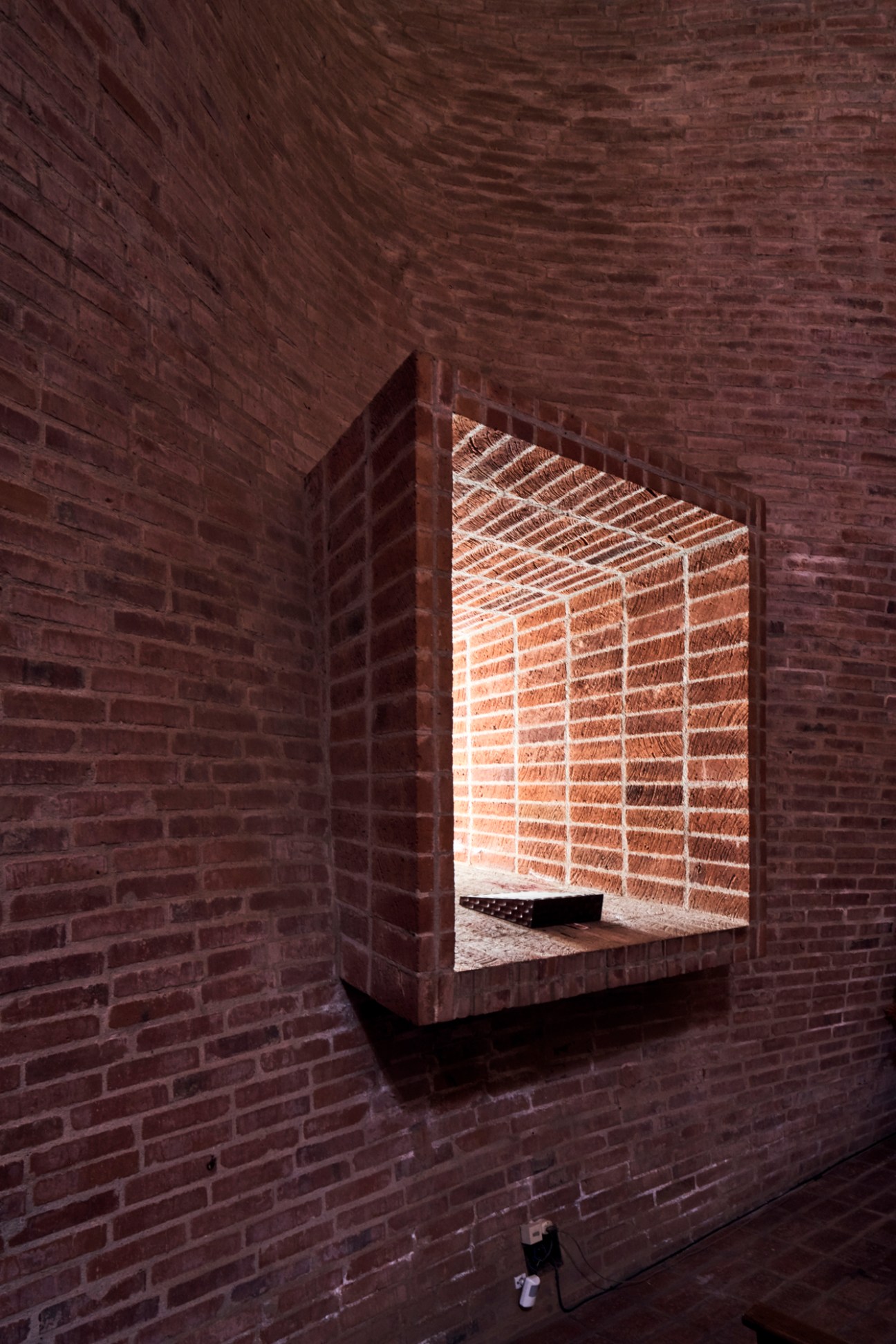 Interior view of Cristo Obrero church showing curved red brick walls with dramatic light filtering through recessed window