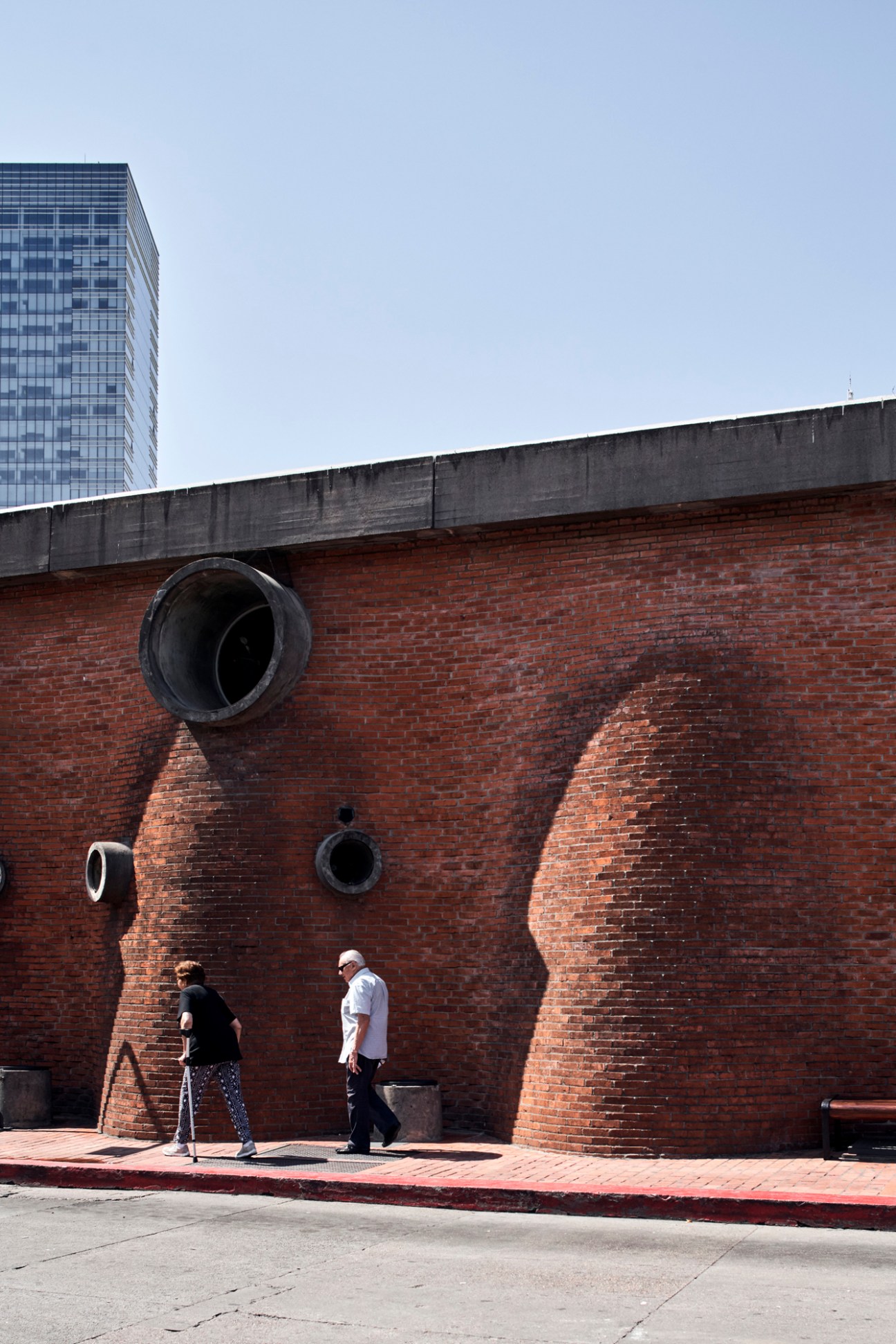 Exterior view of brick building with curved architectural features and circular windows, two people walking past