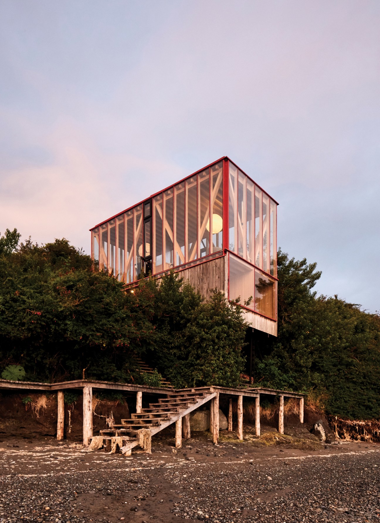 Modern glass and timber cabin elevated on stilts overlooking water with wooden staircase leading up from shoreline