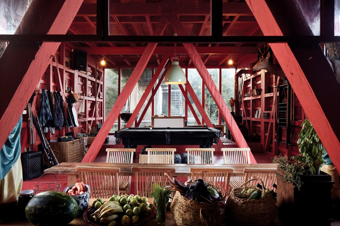 Interior of red timber-framed cabin with pool table, dining area, bookshelves and large windows overlooking forest