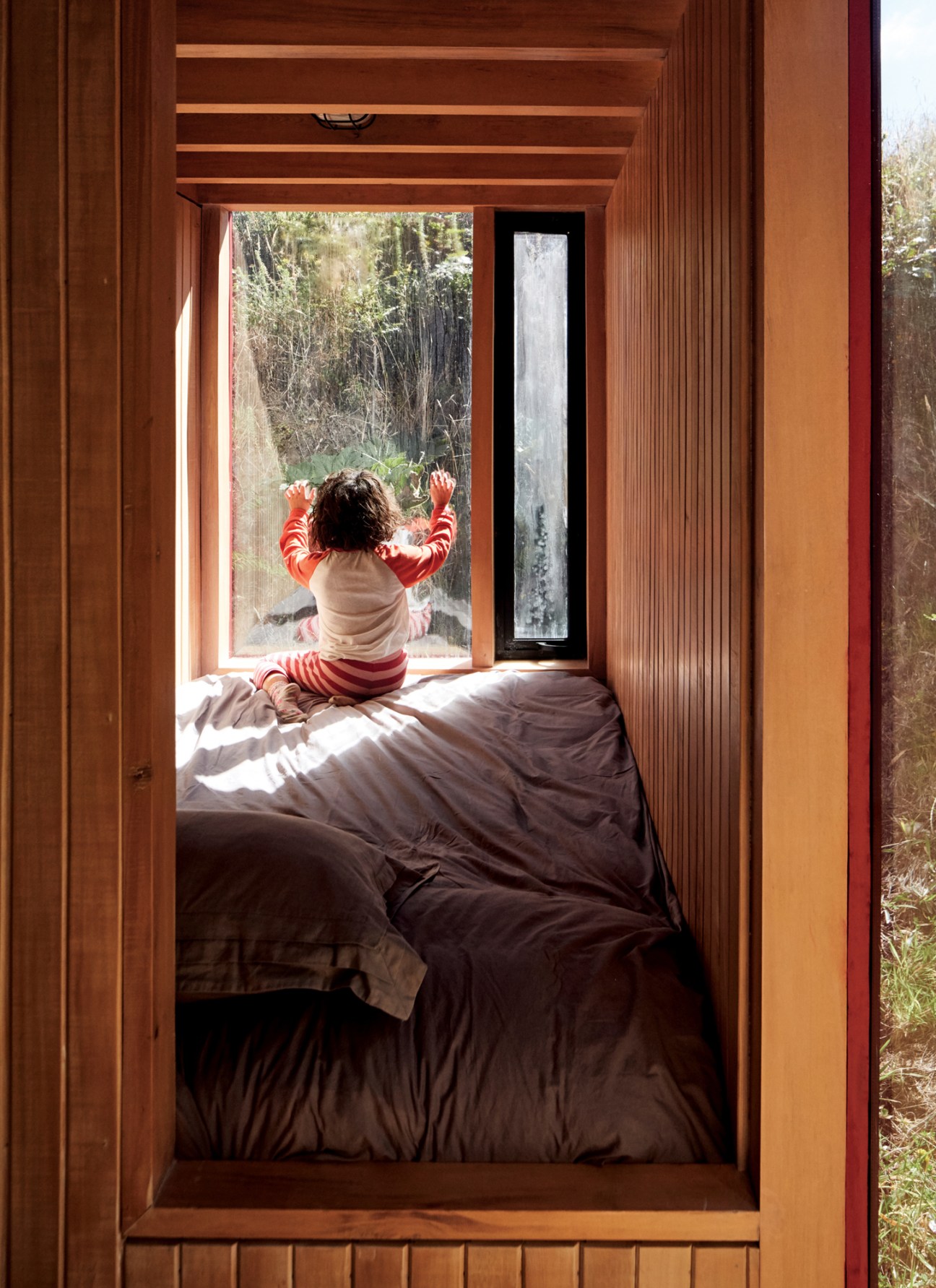 Child in striped red and white pajamas sitting on a bed looking out a large window at forest vegetation