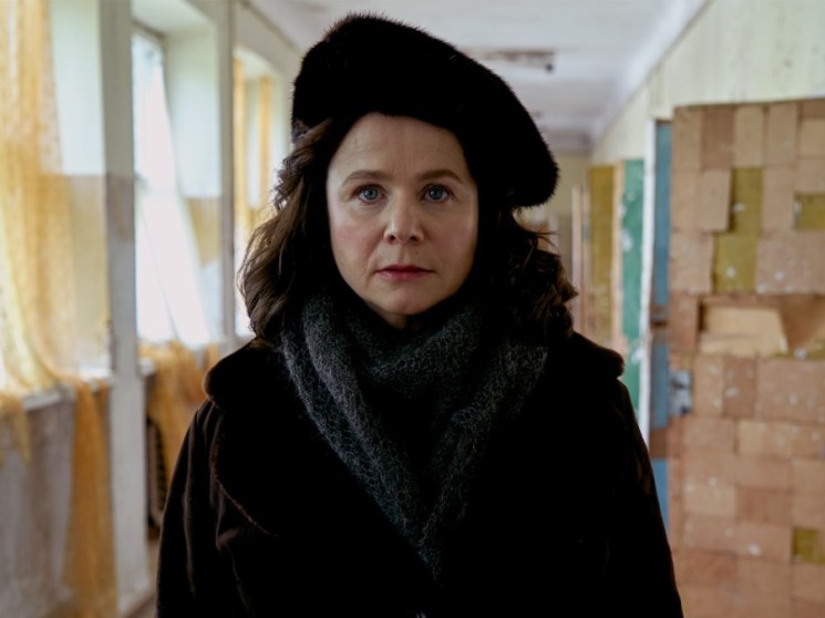 Emily Watson as a Soviet physicist in the Chernobyl mini-series, wearing a black coat and hat in a institutional hallway.