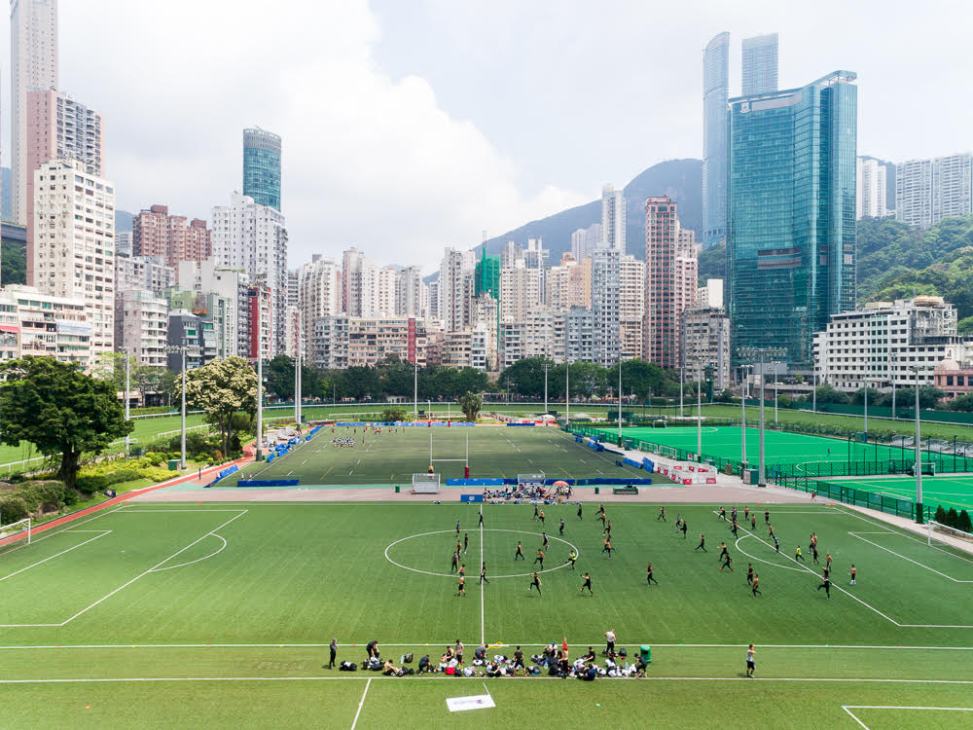 Soccer fields with players and spectators overlooking Hong Kong's dense urban skyline with mountains.