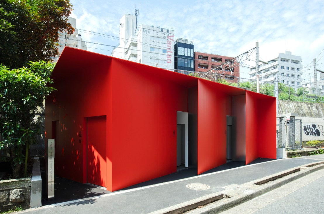 Modern red public toilet building with three entrance bays on a Tokyo street, surrounded by urban buildings