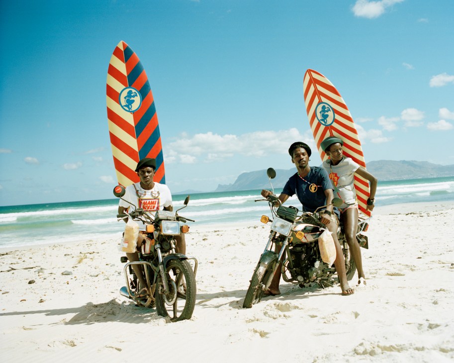 Three men with motorcycles and surfboards on a beach with ocean waves and mountains in background