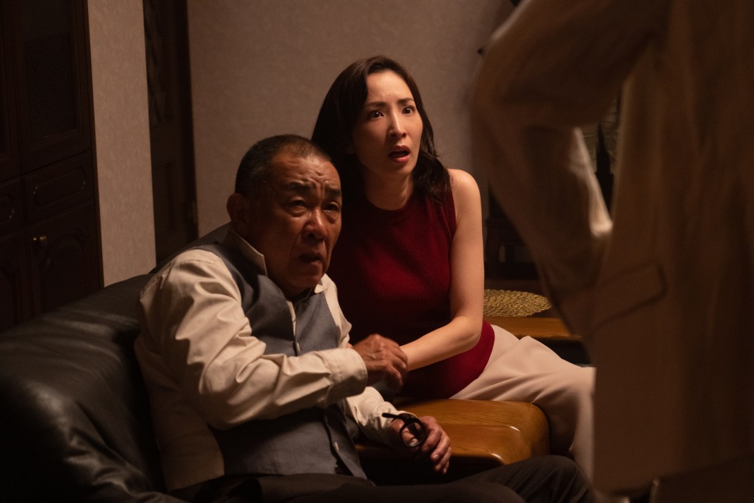 Two people sitting together on a bed in a dimly lit room, looking concerned or distressed.