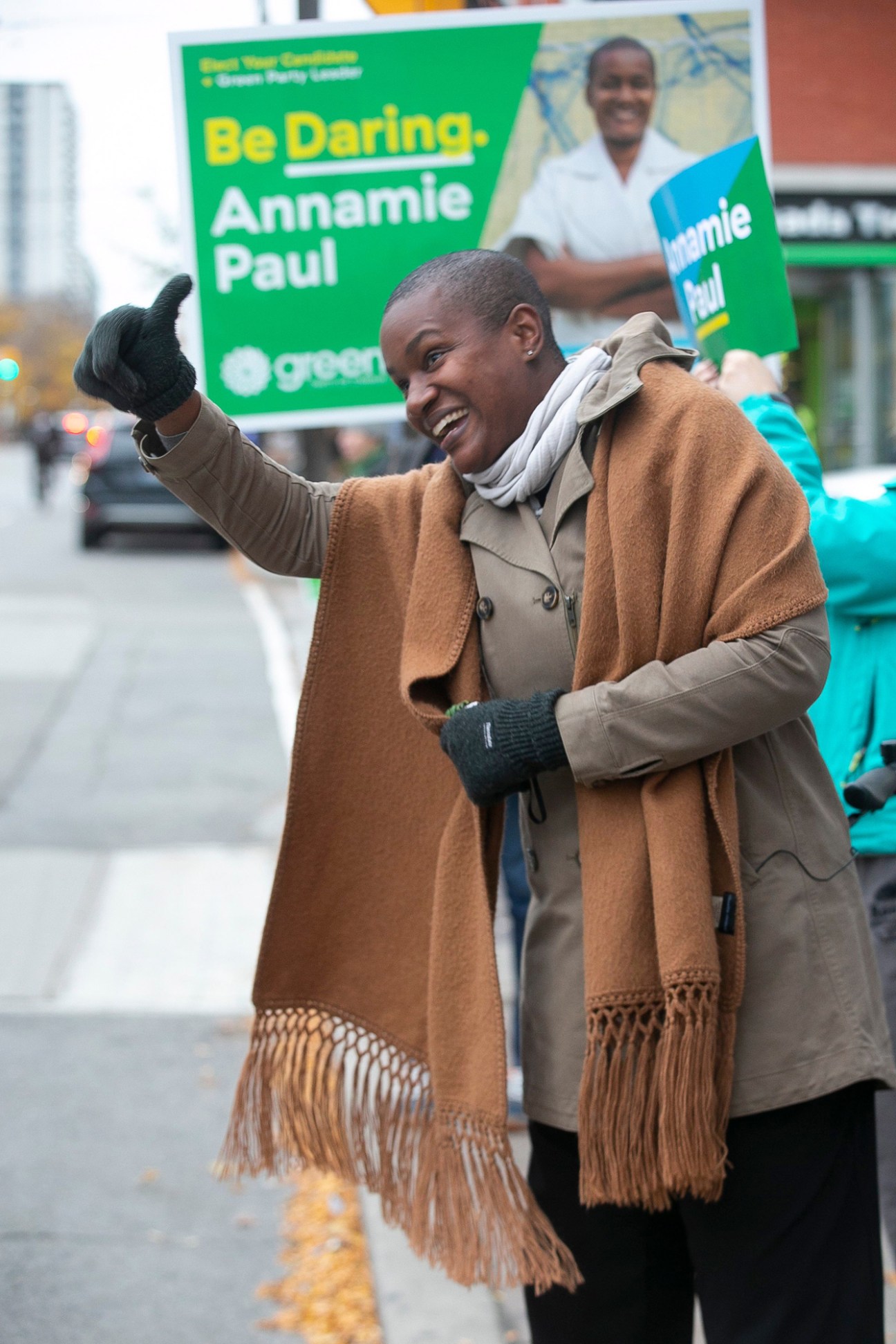 Annamie Paul giving thumbs up while campaigning, wearing brown coat and scarf with Green Party signs behind her