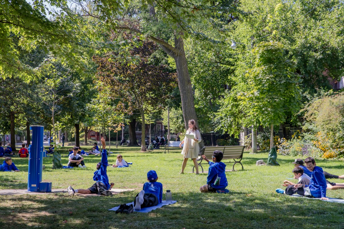 Children in blue jackets sitting on grass in a park while an adult in white leads an outdoor class