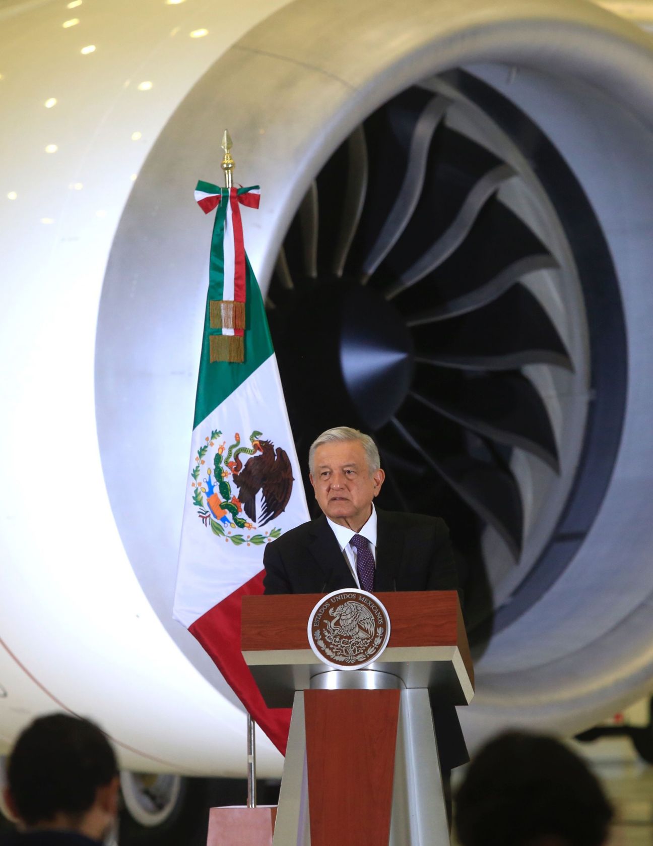 Mexican president speaking at podium with Mexican flag and large aircraft engine in background