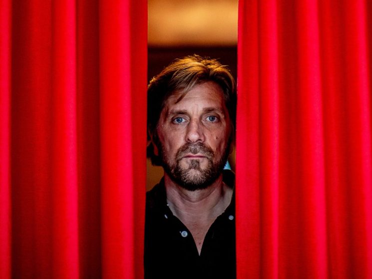Swedish film director Ruben Östlund peering through red curtains with chandelier visible in background