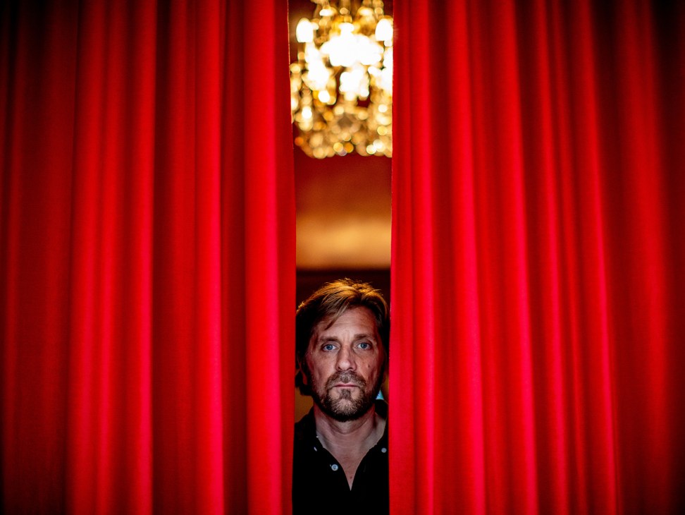 Swedish film director Ruben Östlund peering through red curtains with chandelier visible in background