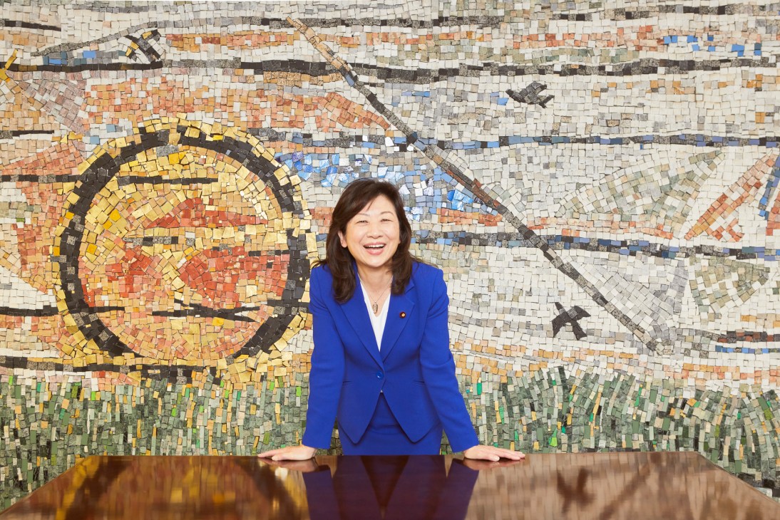 Seiko Noda smiling in a bright blue blazer standing behind a wooden table with an ornate mosaic wall backdrop