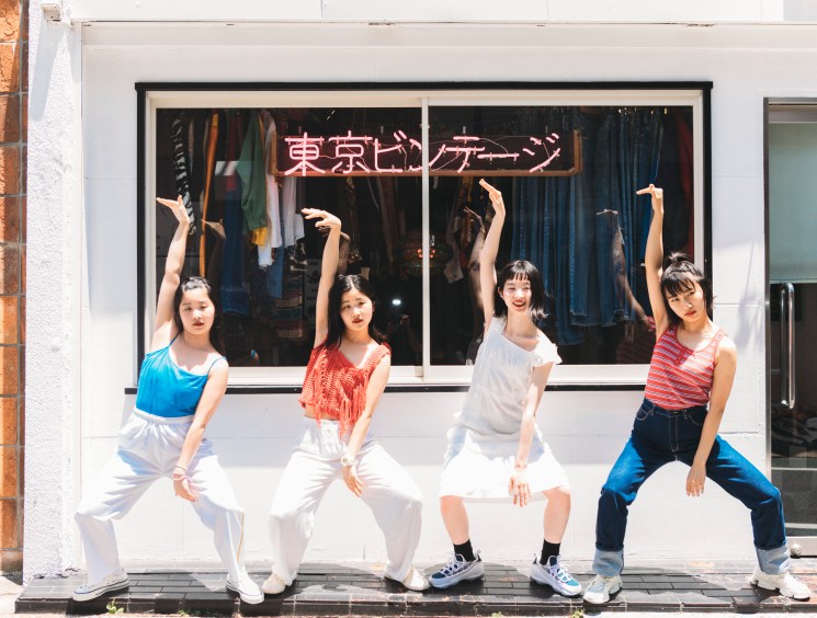 Four young women pose playfully outside a Tokyo vintage clothing shop with neon Japanese signage.