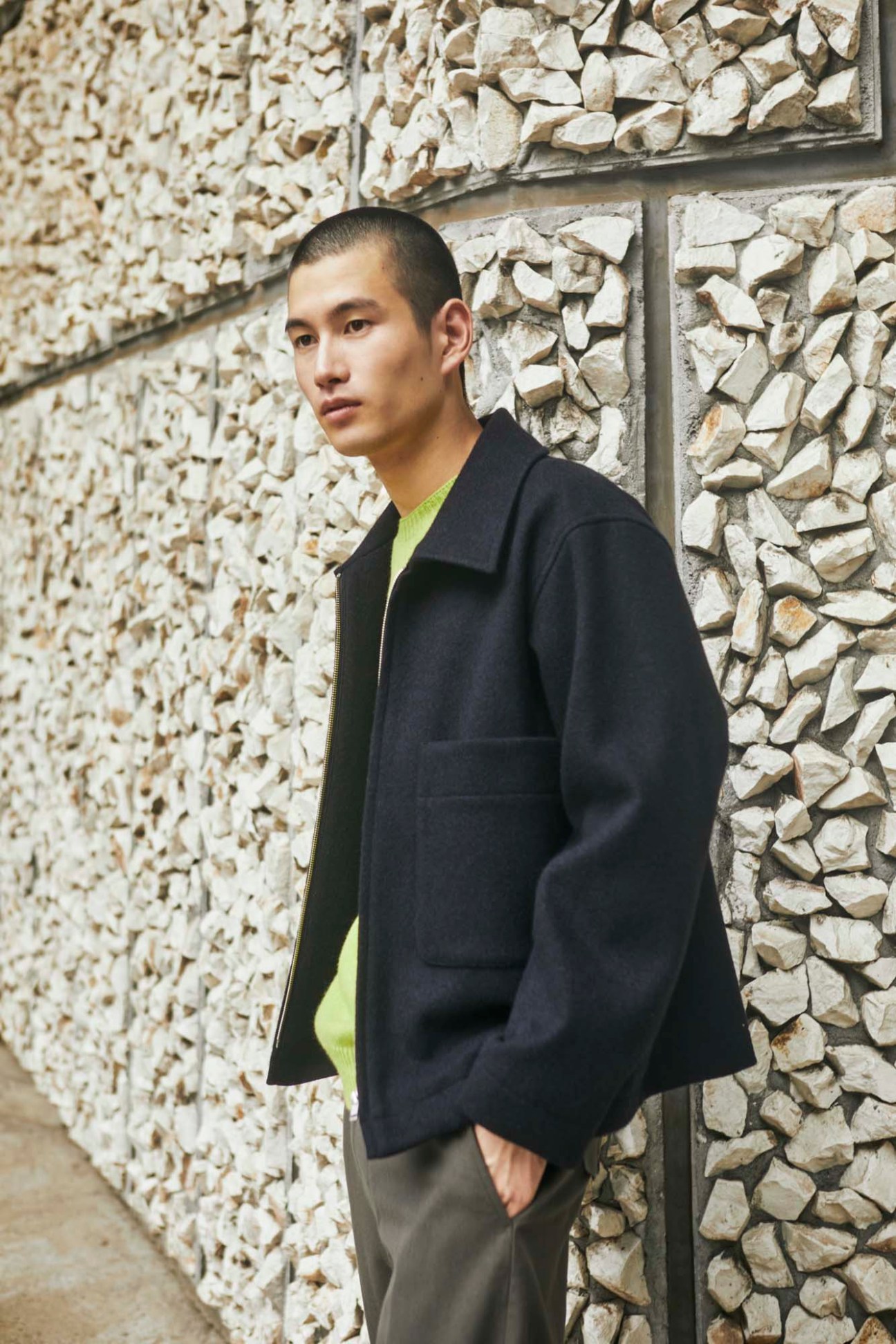 Young Asian man in black jacket and yellow jumper standing against textured stone wall background