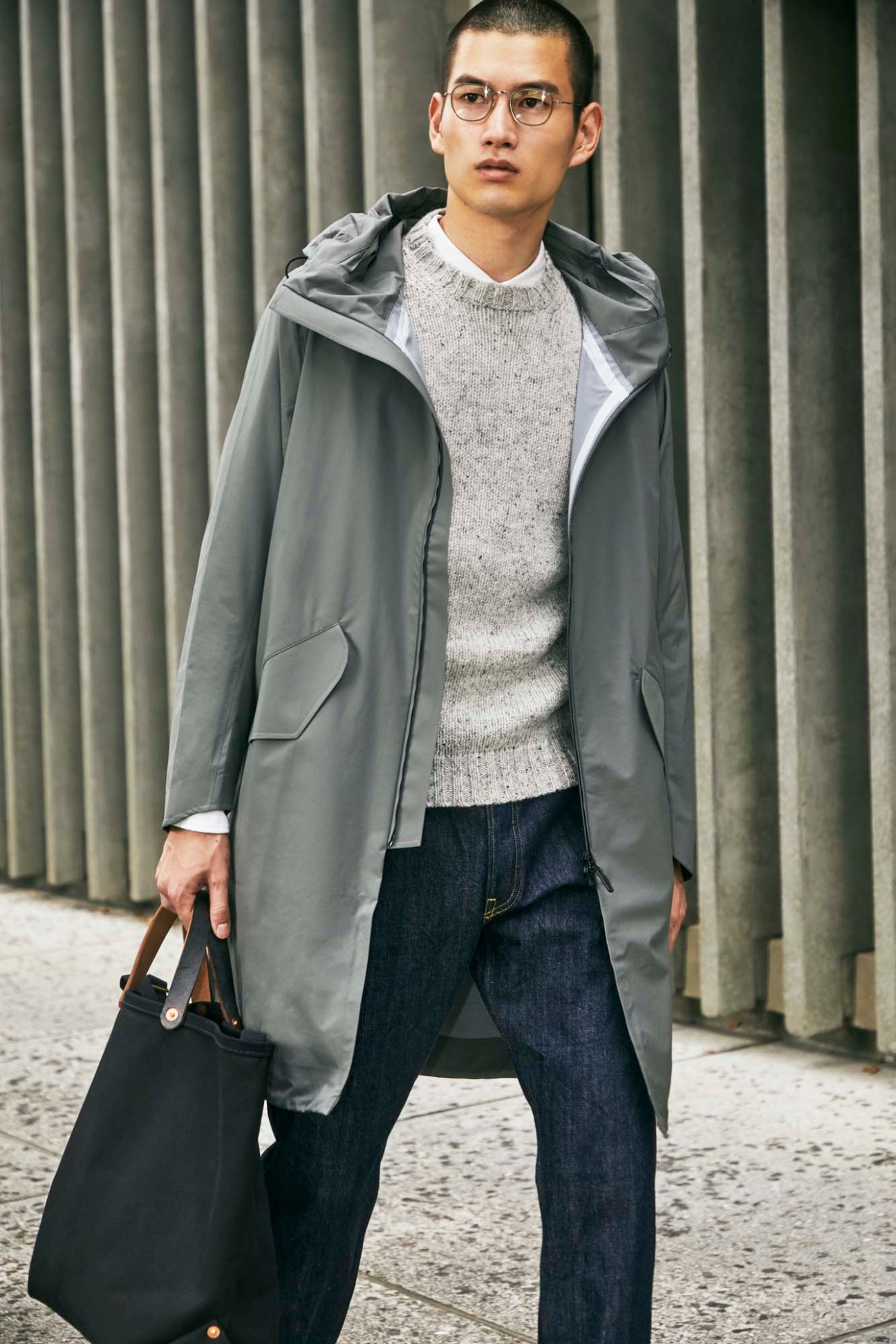 Young man in glasses wearing grey hooded coat over cream sweater, carrying black bag on urban street