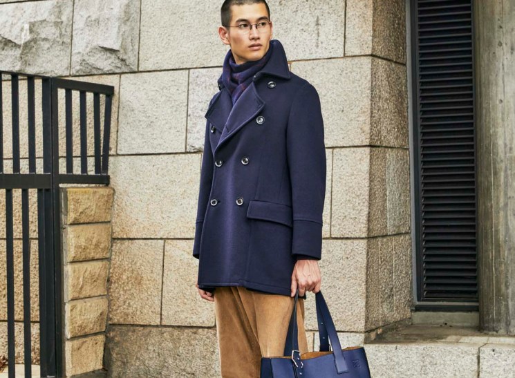 Young man in navy peacoat and brown trousers holds blue leather bag, standing against stone building wall